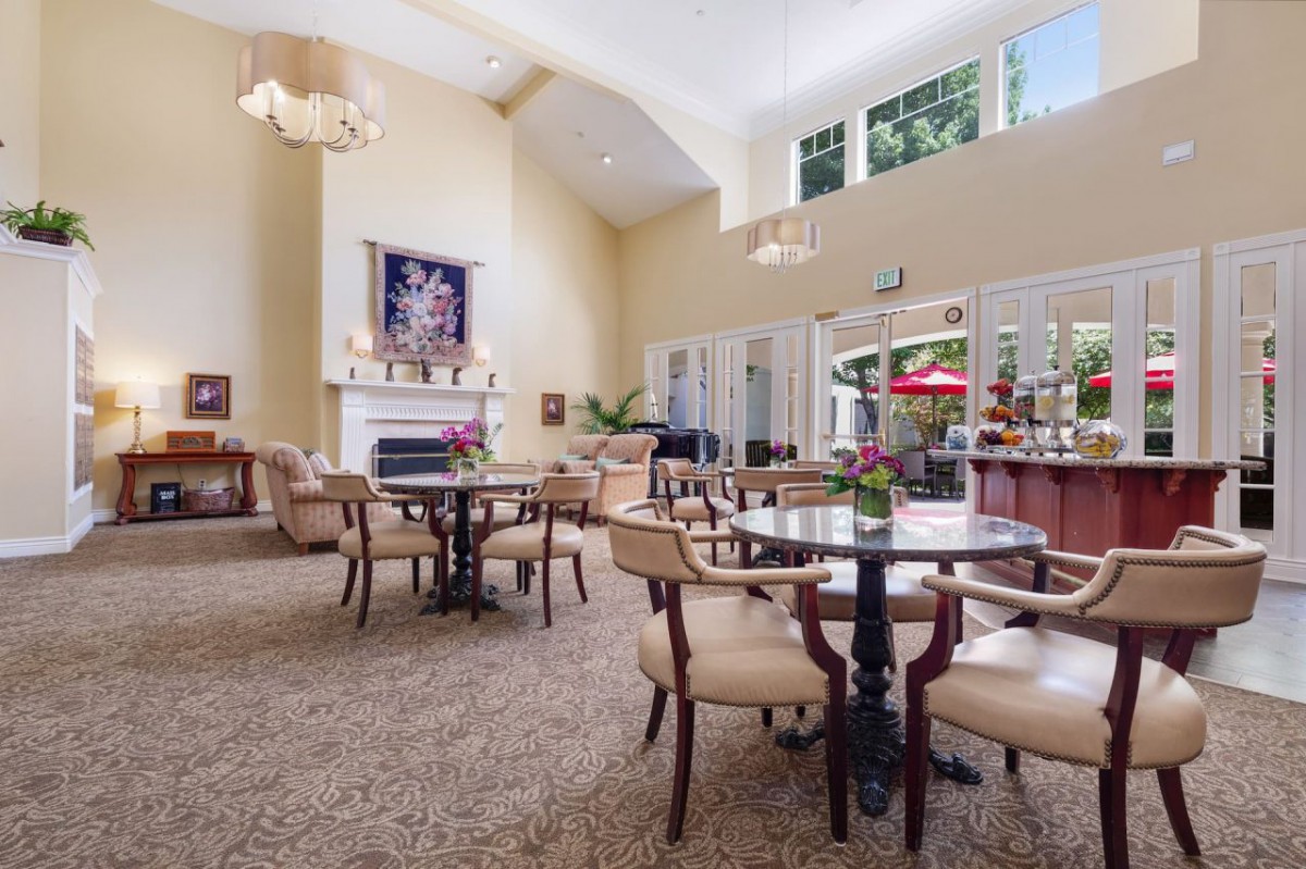 A spacious and elegantly decorated lounge area featuring multiple round tables with chairs, a fireplace, and large windows allowing natural light to flood the room.