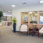 A well-lit lobby featuring comfortable seating and a small table, with large windows providing a view of the adjoining room.