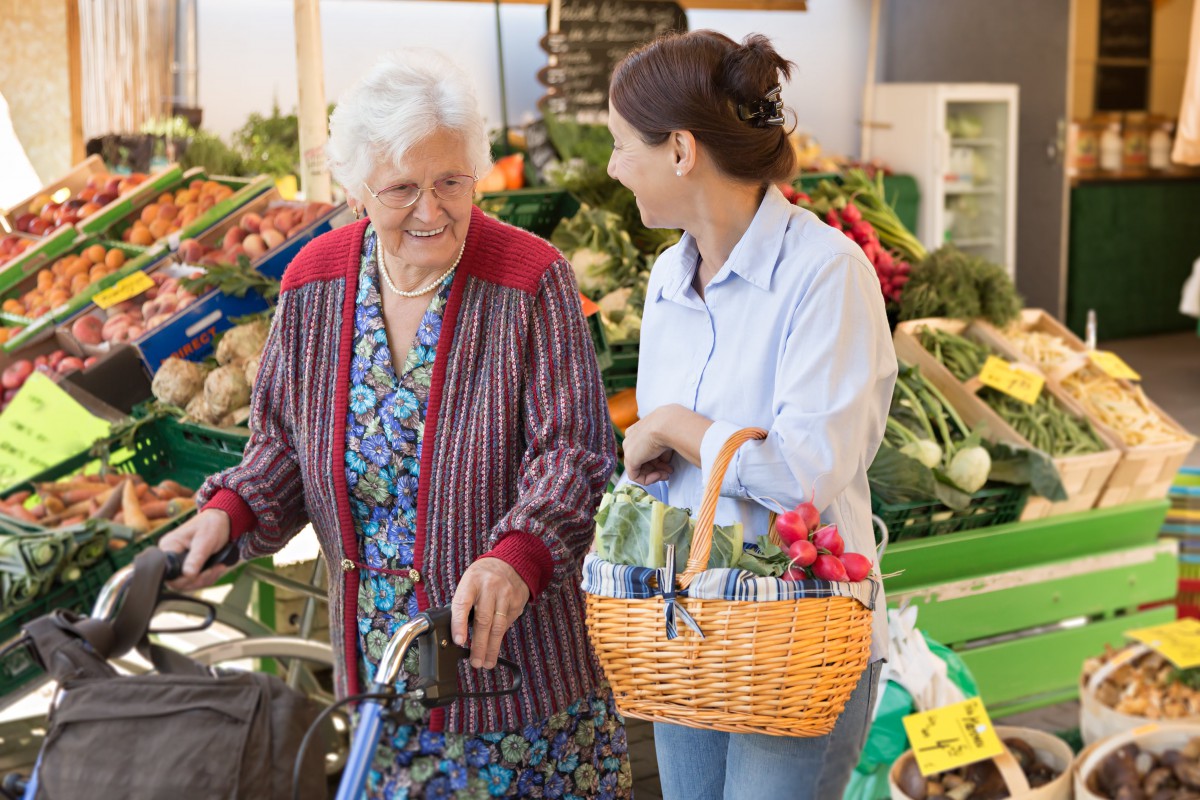 A young woman assists an elderly woman with grocery shopping at a market.