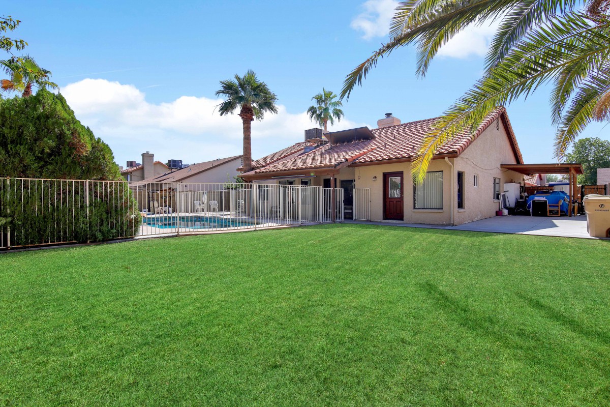 A beautiful house with a spacious backyard featuring a green lawn, swimming pool, and palm trees under a sunny sky.