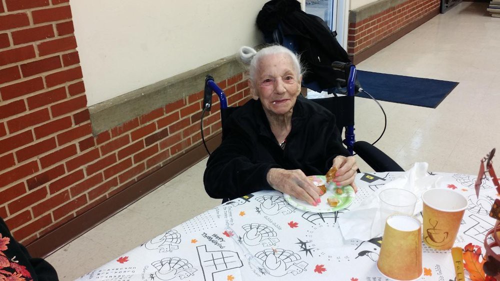 An elderly woman sits in a wheelchair at a table with coloring pages, enjoying a snack.