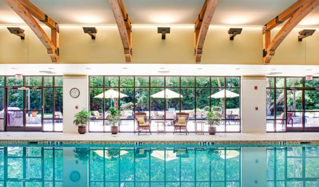 A serene indoor swimming pool with a view of outdoor seating and umbrellas through large glass windows.