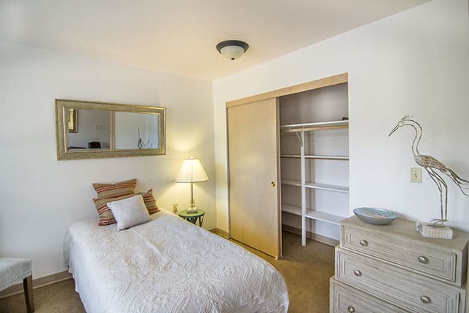 A minimalist bedroom with a single bed, a lamp on a side table, an open closet, and a dresser.