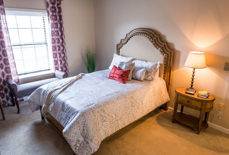 A cozy bedroom featuring a neatly made bed with decorative pillows, a bedside lamp on a wooden nightstand, and natural light coming through the window with patterned curtains.