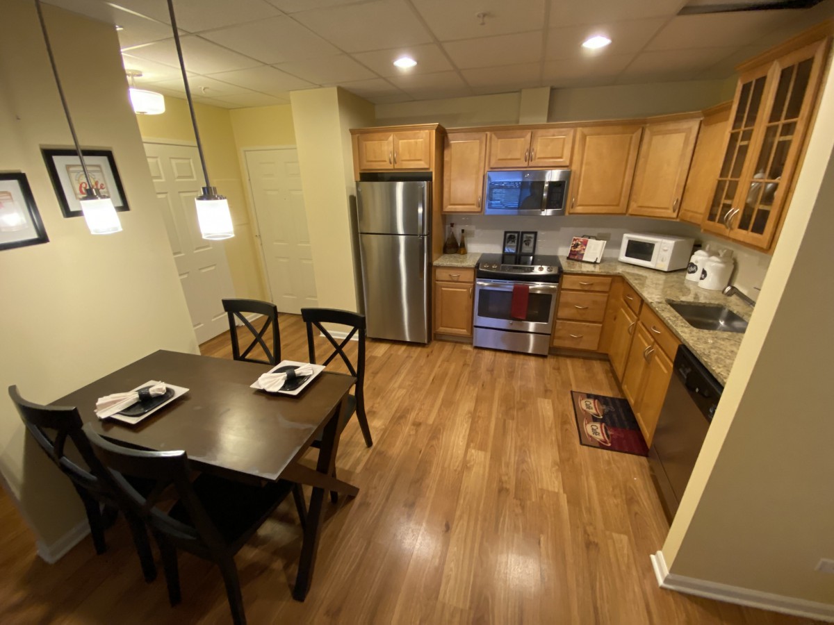 A modern kitchen with wooden flooring, featuring a dining table and chairs, stainless steel appliances, and pendant lighting.