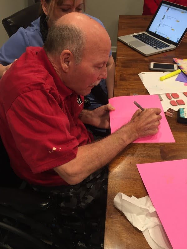 A person in a red shirt writing on pink paper at a table with stationery and a laptop.