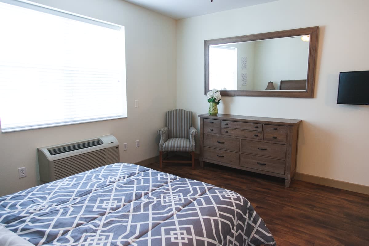 A cozy bedroom with a patterned bedspread, wooden dresser with a mirror, and an upholstered armchair.