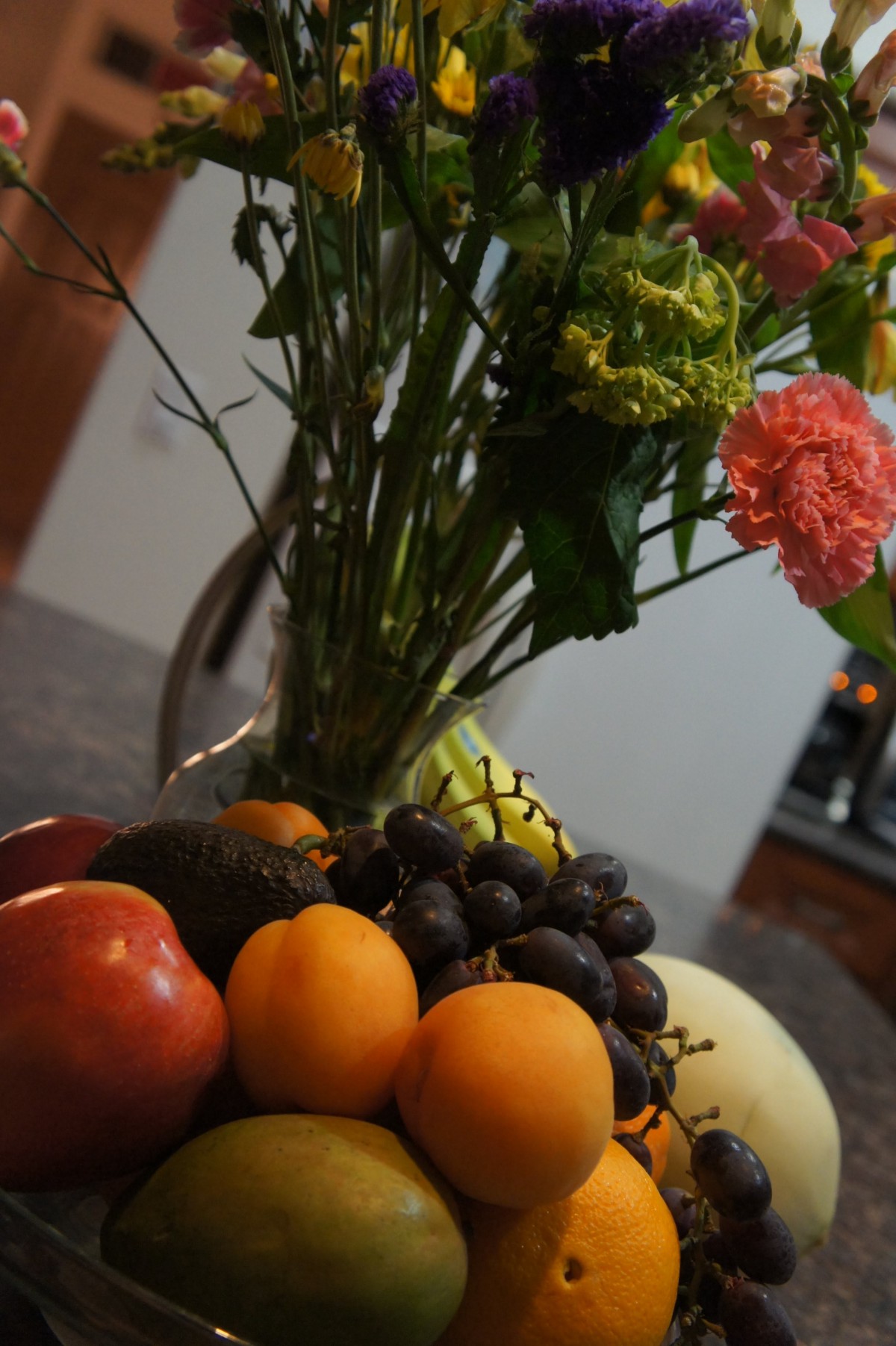 A fruit bowl filled with various fruits next to a bouquet of flowers on a table.