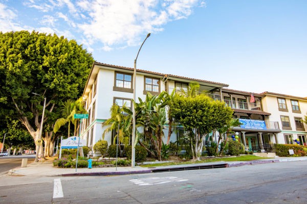 A view of an apartment building on a sunny day, surrounded by trees along a street corner.