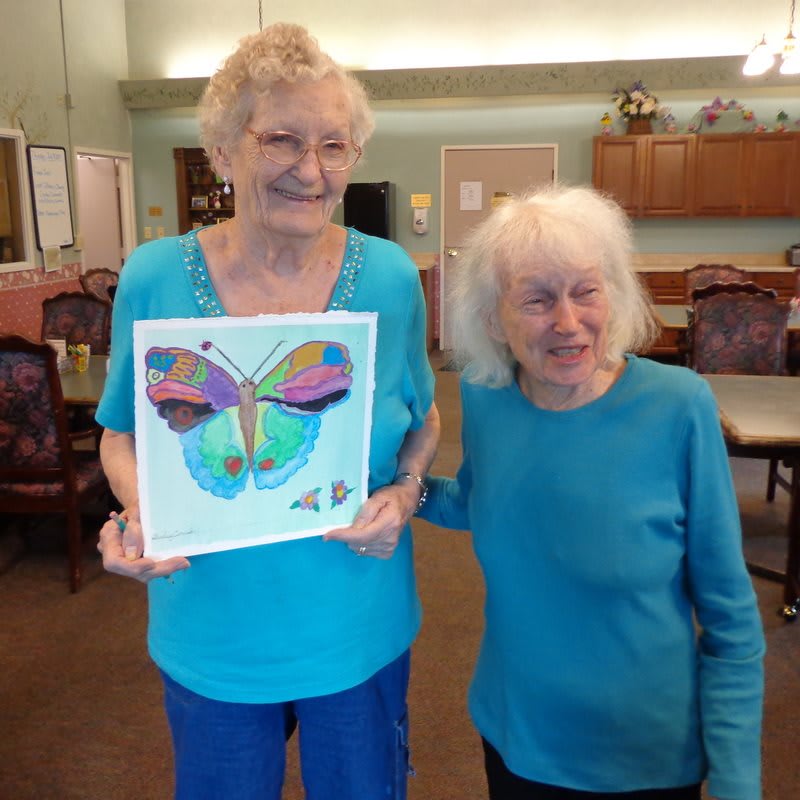 Two elderly women smiling, one holding colorful butterfly artwork indoors.