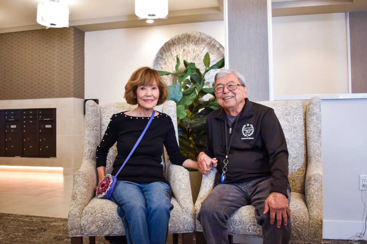 An elderly couple sitting indoors, holding hands and smiling.