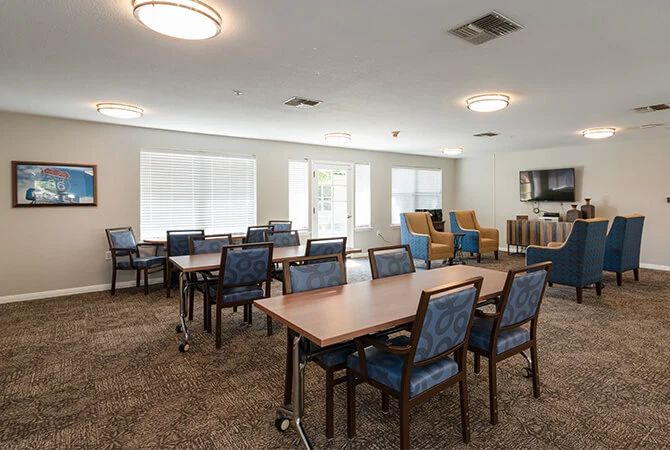 A well-lit meeting room with multiple tables and chairs, carpeted flooring, and large windows.