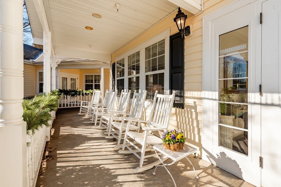A sunny porch featuring several white rocking chairs lined up, with a flower pot adding a touch of color.