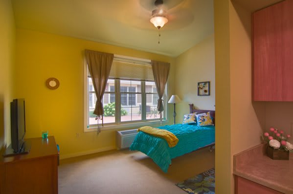 A cozy bedroom with yellow walls and a turquoise bedspread, featuring large windows with curtains and a ceiling fan.