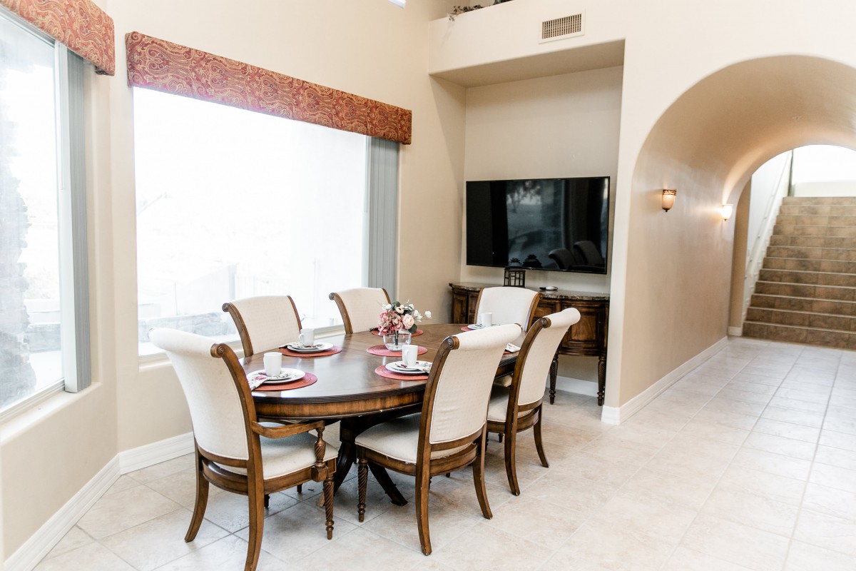 A well-lit dining area with a wooden table and chairs, featuring a large window and a television mounted on the wall.