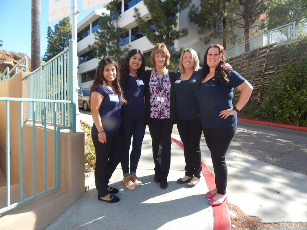 A group of five women smiling and standing together outdoors in casual clothing.