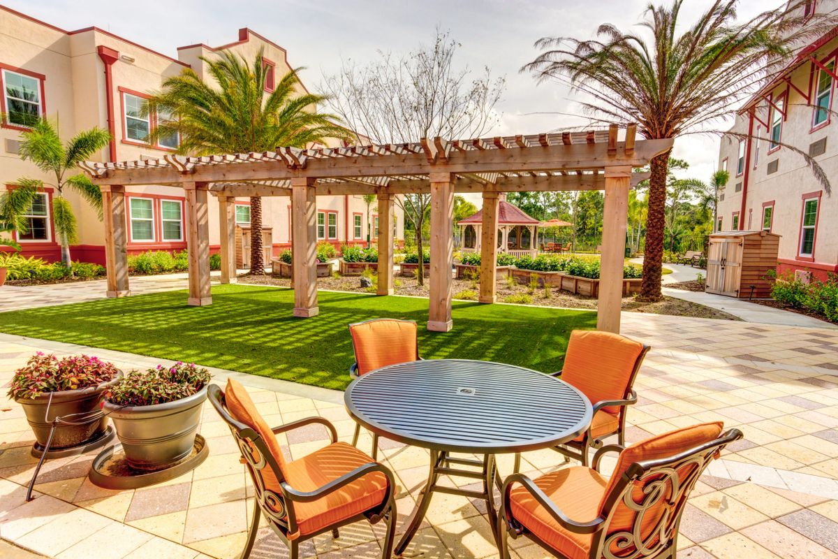 A sunny patio area with a table and orange chairs, surrounded by a pergola and palm trees.