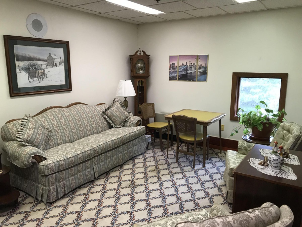 A cozy vintage-style living room with a patterned sofa, a small dining table, a grandfather clock, and indoor plants.