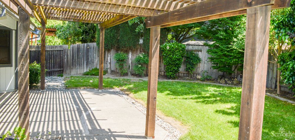 A backyard with a wooden pergola casting shadows on the patio, surrounded by a green lawn and a wooden fence.
