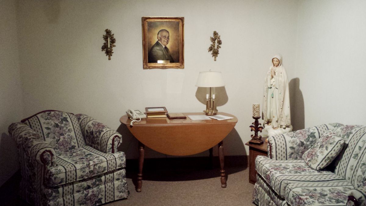 A cozy living room with a central wooden table, flanked by two armchairs. A portrait hangs on the wall above the table, accompanied by decorative elements such as a lamp and a statue.