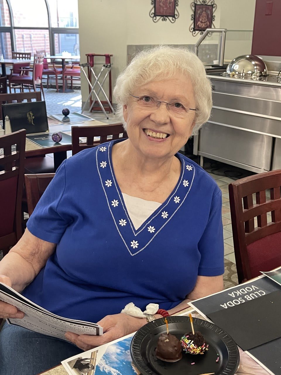An elderly woman smiling while seated at a dining table indoors.