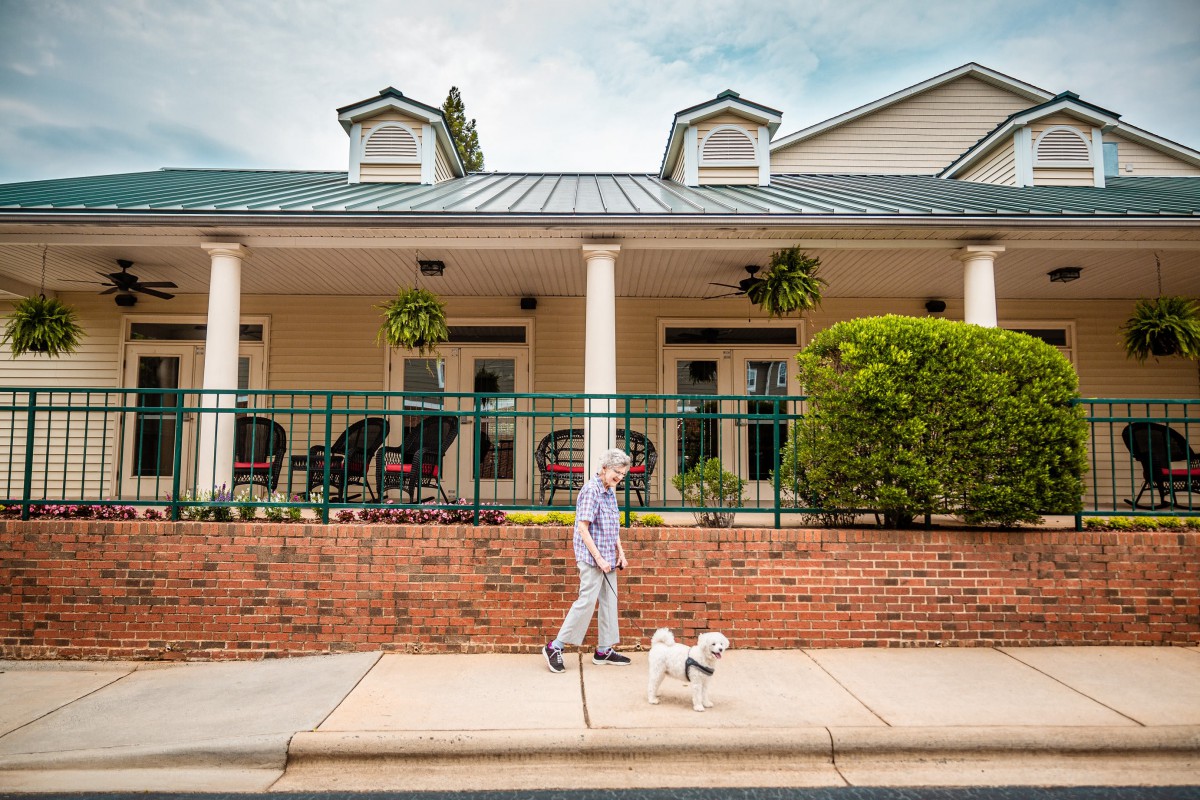 A woman walks her dog on a sidewalk past a house with a porch and hanging plants.