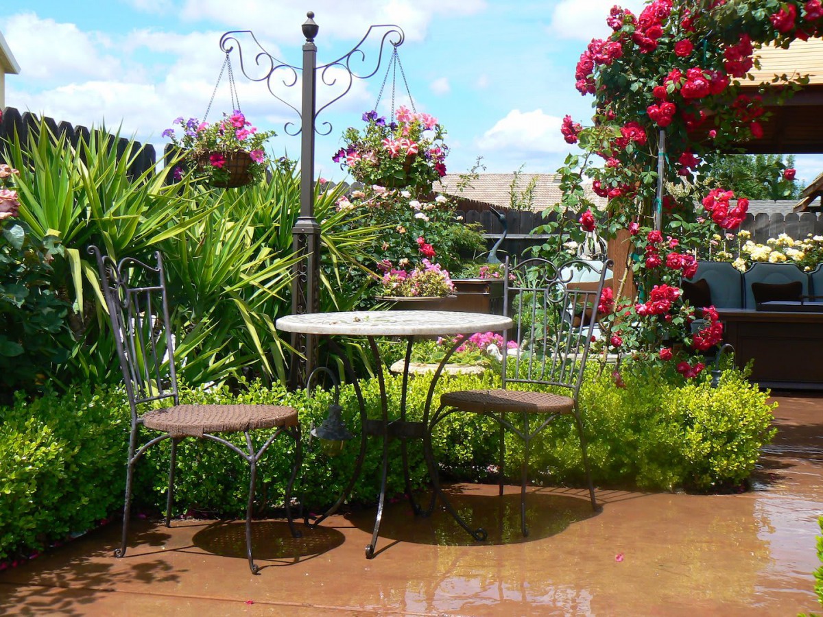 A charming garden patio with a table and two chairs surrounded by lush greenery and blooming roses.
