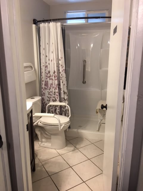 A small bathroom with a toilet featuring support rails, a shower area with a grab bar, and decorative shower curtain.