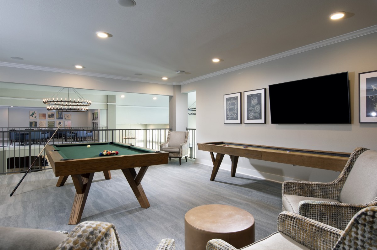 A modern game room featuring a pool table, shuffleboard table, and seating area with a wall-mounted television.
