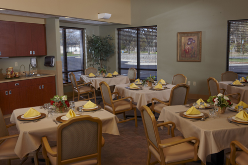 A well-furnished dining room set for a meal, featuring elegantly arranged tables with yellow napkins and a pleasant view outside.