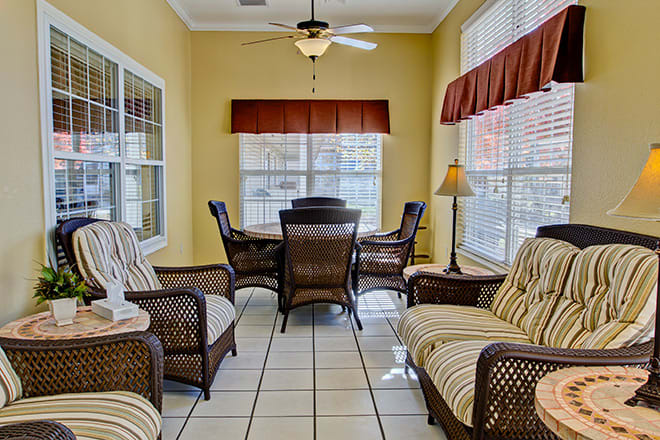 A cozy sunroom with rattan furniture and a small dining table, featuring large windows and a ceiling fan.