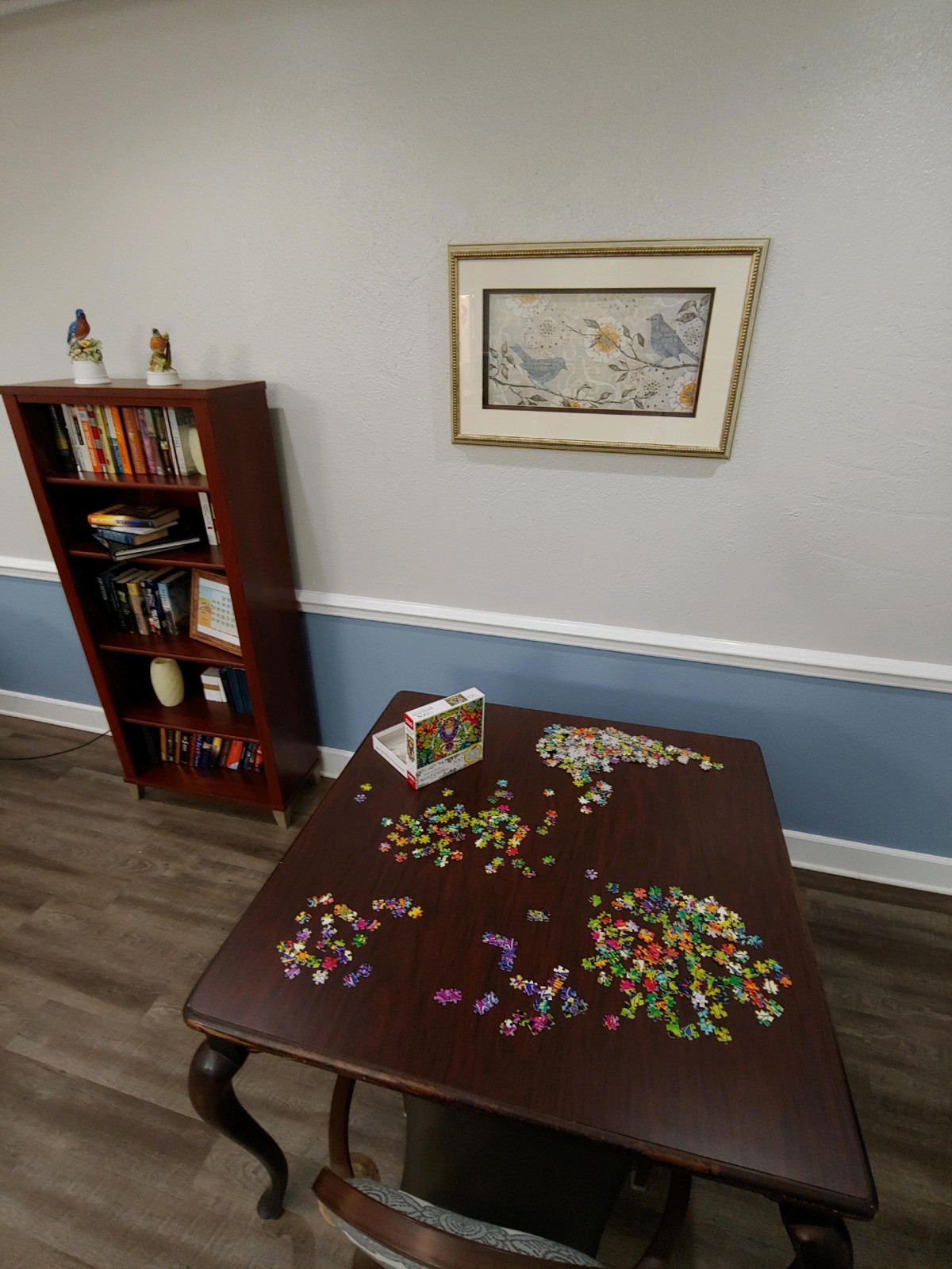 A room with a partially completed puzzle on a table, a bookshelf with books and decorations, and a painting on the wall.