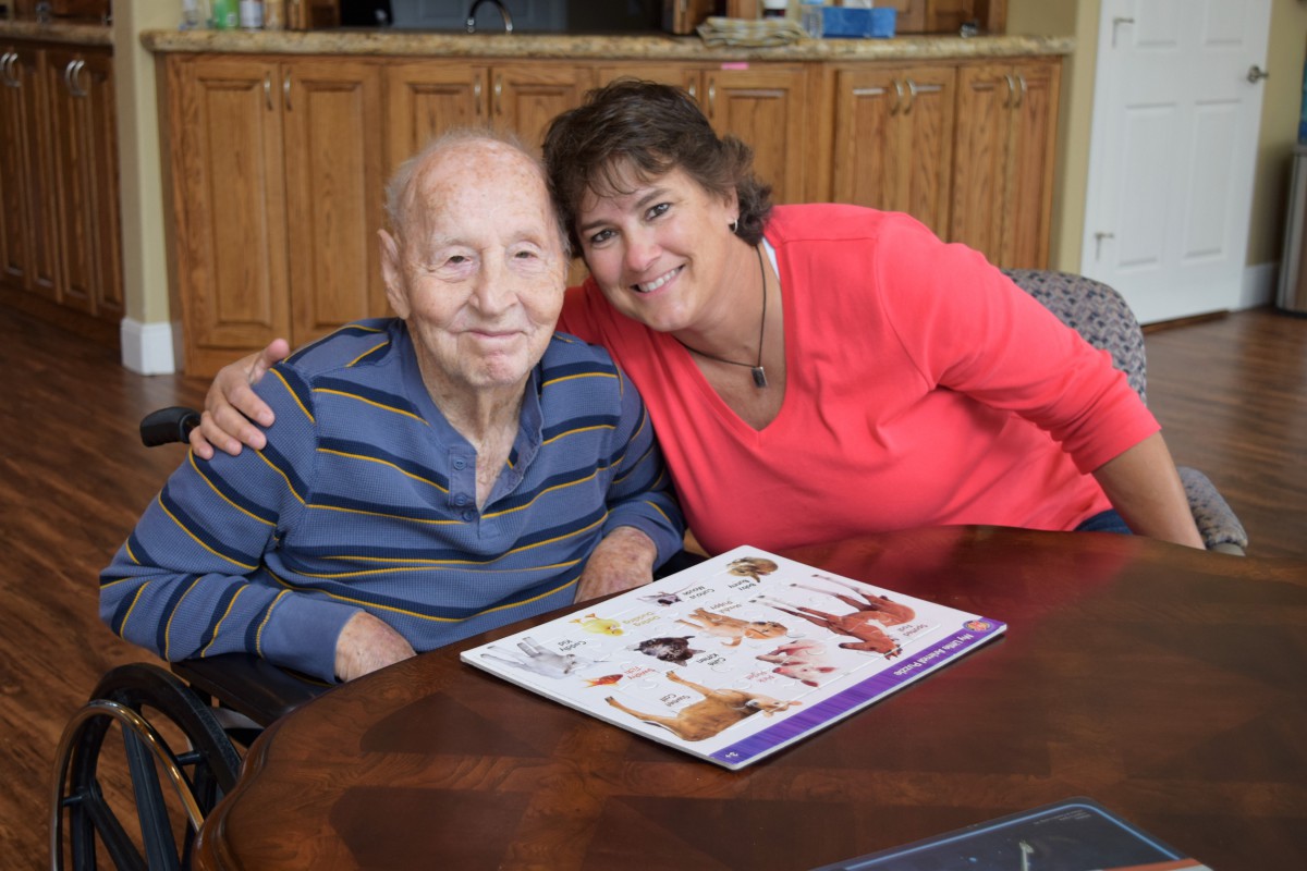 A woman and an elderly man are sitting together at a table with a booklet in front of them, smiling warmly.