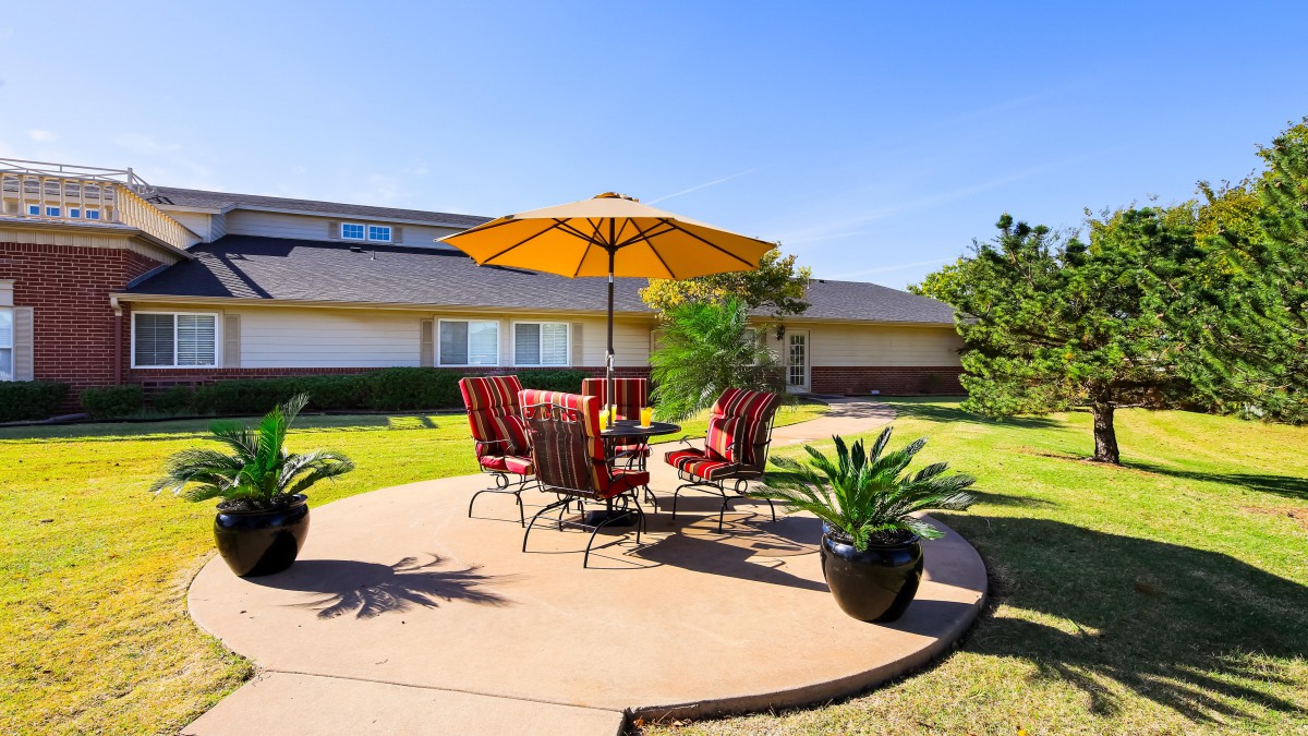 A sunny backyard patio with a table, chairs, and an umbrella surrounded by a well-maintained lawn and potted plants.