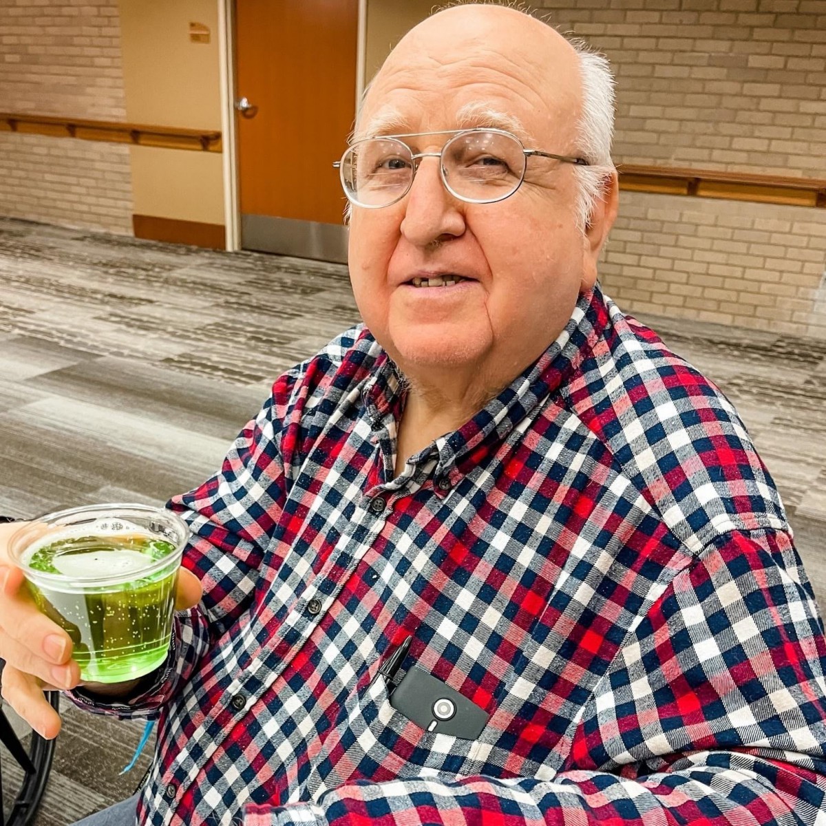 An elderly man wearing a plaid shirt holds a drink while sitting outdoors.