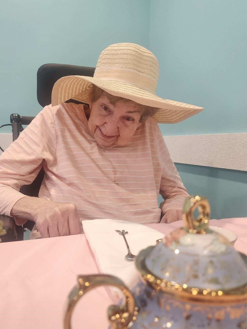 An elderly woman wearing a wide-brimmed hat, sitting in a wheelchair at a table with writing materials.