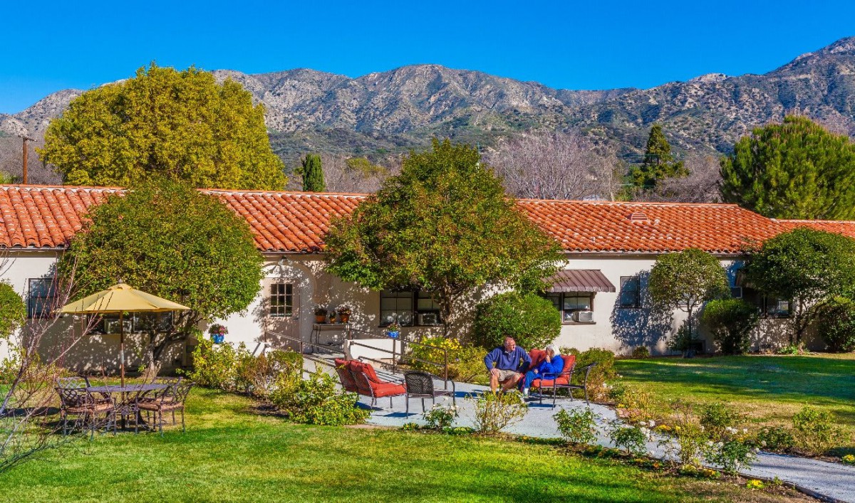 A charming house with a red-tiled roof is set against a backdrop of mountains, surrounded by lush gardens and trees, with colorful patio furniture in the foreground.