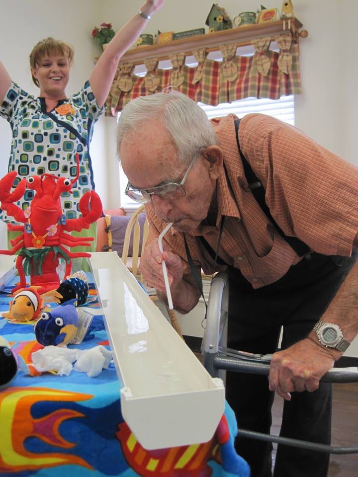 An elderly man participates in a fun indoor game using a straw, while another person cheers in the background.