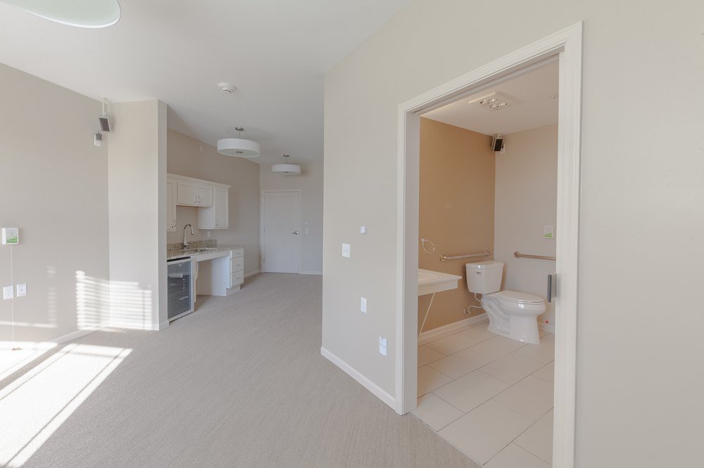 A modern apartment interior featuring a compact kitchen and adjacent bathroom with light-colored walls.