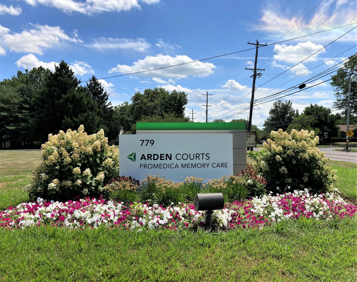 A roadside sign for Arden Courts Promedica Memory Care surrounded by well-maintained landscaping and flowers.