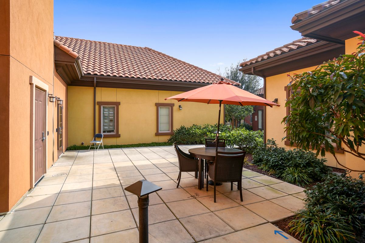 A serene outdoor patio with tiled flooring, featuring a table and chairs under an umbrella, surrounded by a garden and yellow stucco buildings.