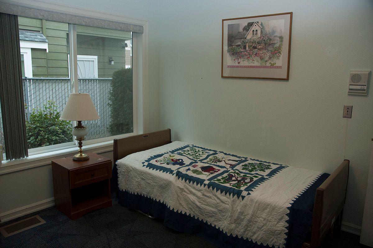 A simple bedroom with a single bed, nightstand, and lamp next to a large window.