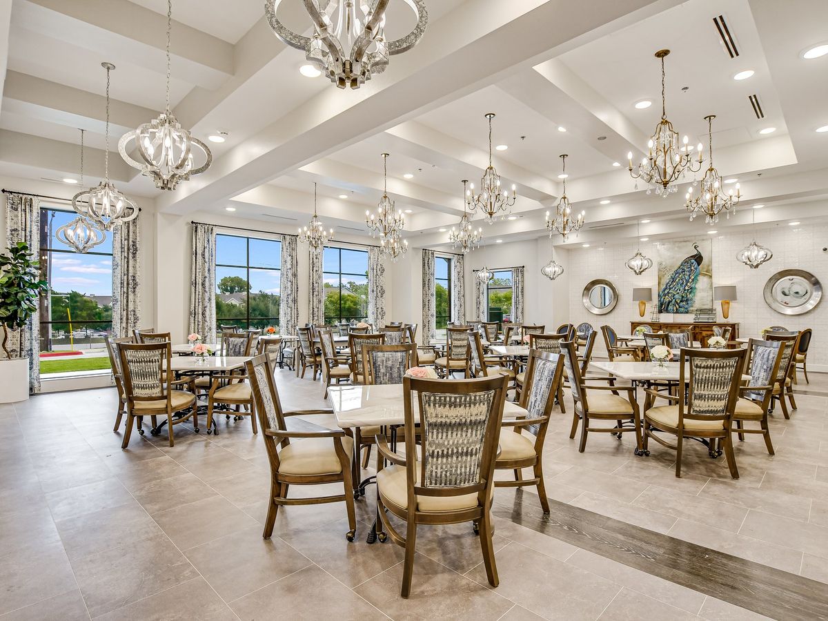 A spacious and elegantly designed dining room with chandeliers, featuring multiple tables and chairs under bright lighting.