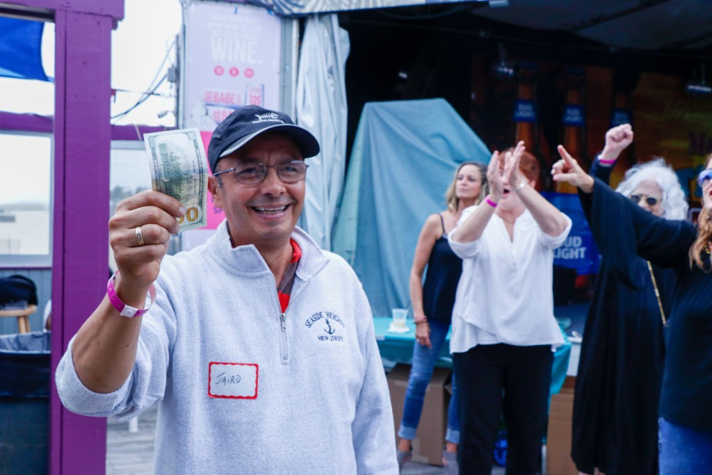 A man holding up money, smiling at an outdoor event where people are cheering in the background.