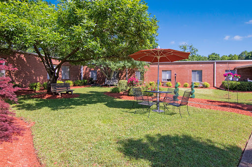A serene outdoor garden space featuring a seating area under an umbrella, surrounded by lush greenery and a charming brick building.