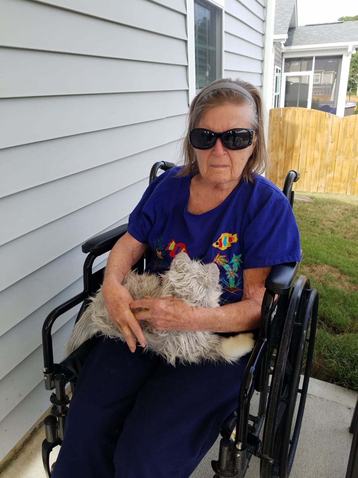 An elderly woman wearing sunglasses is sitting in a wheelchair outside, holding a cat.