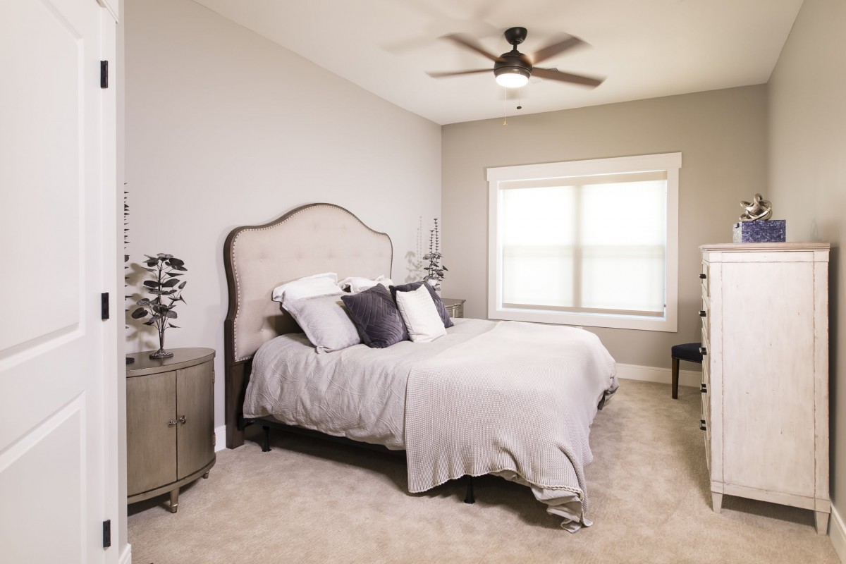 A modern bedroom with neutral colors featuring a comfortable bed, ceiling fan, and a carpeted floor.