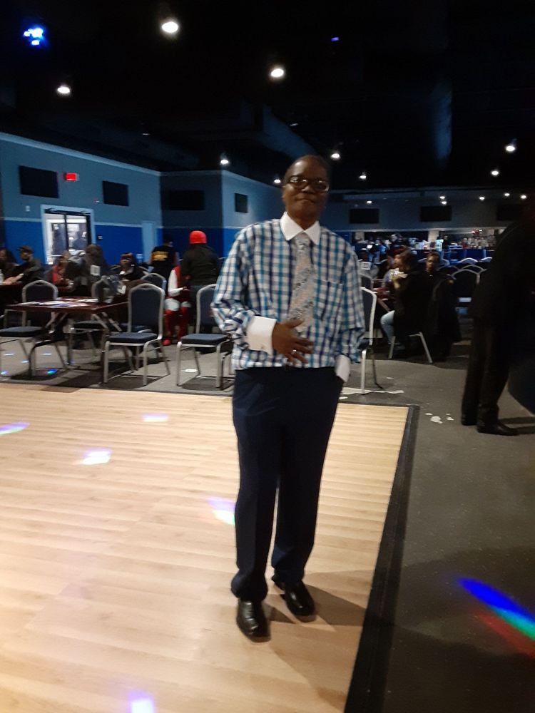 A person standing in a venue with tables and chairs, dressed in a plaid shirt and dark trousers.