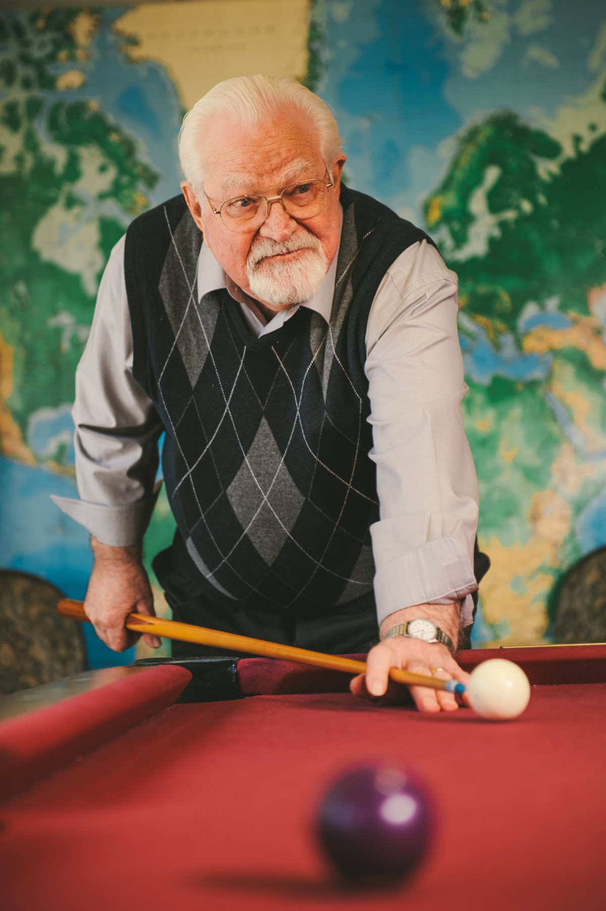 An elderly man playing billiards on a red pool table with a world map in the background.