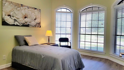 A cozy bedroom featuring a single bed, a lamp, and two large windows that allow natural light to fill the space.